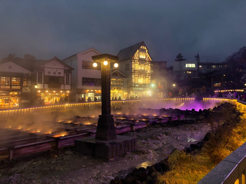 The yubatake at night. All the buildings are lit up, and the yubatake itself is lit with purple and yellow lights that rise up through the fog.