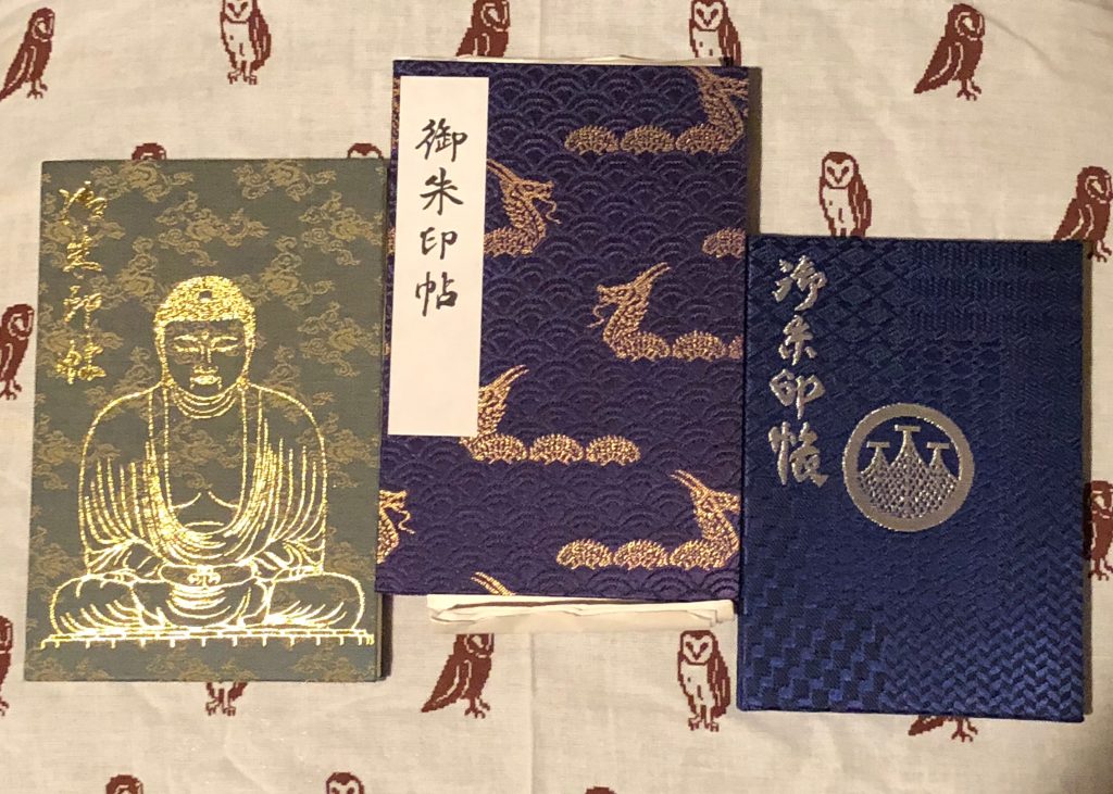 Three different goshuin books. The one on the far left is green with a golden Buddha statue stamped on it -- it's clearly from Kamakura. The one in the center is blue with golden dragons. The one on the right is the simplest design, blue with three towers in silver foil.