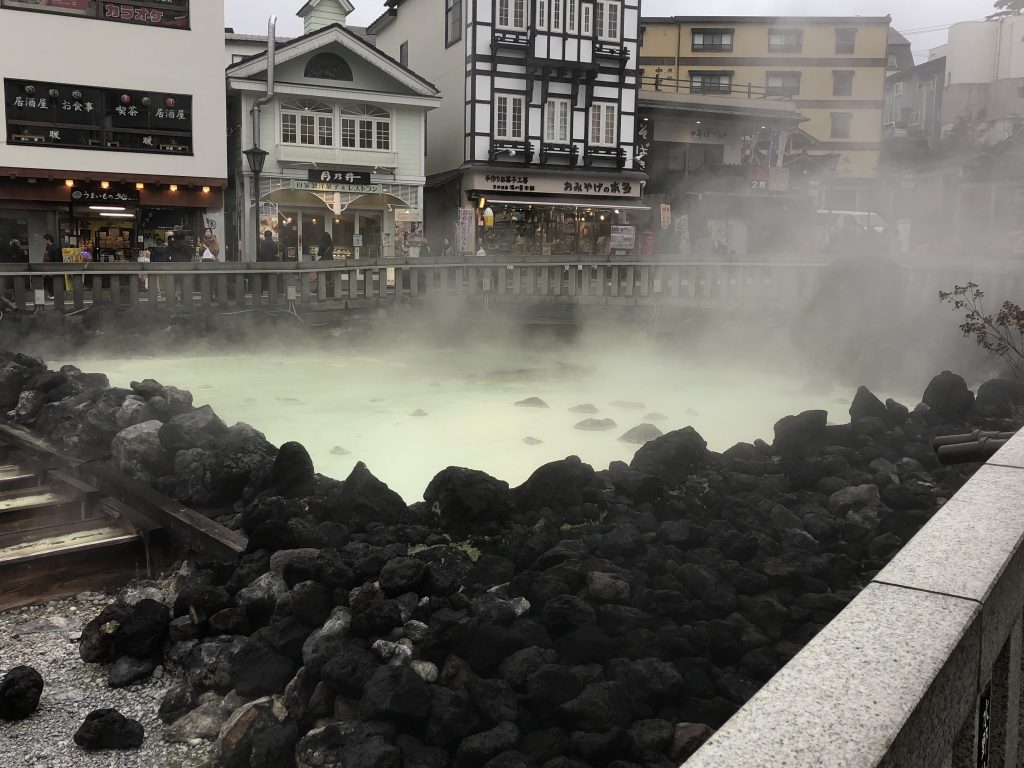 A hot spring shrouded in immense amounts of steam. Through the steam, you can see a pool of water that is a milky green. The pool is surrounded with black rocks. The town shops can be seen in the background.