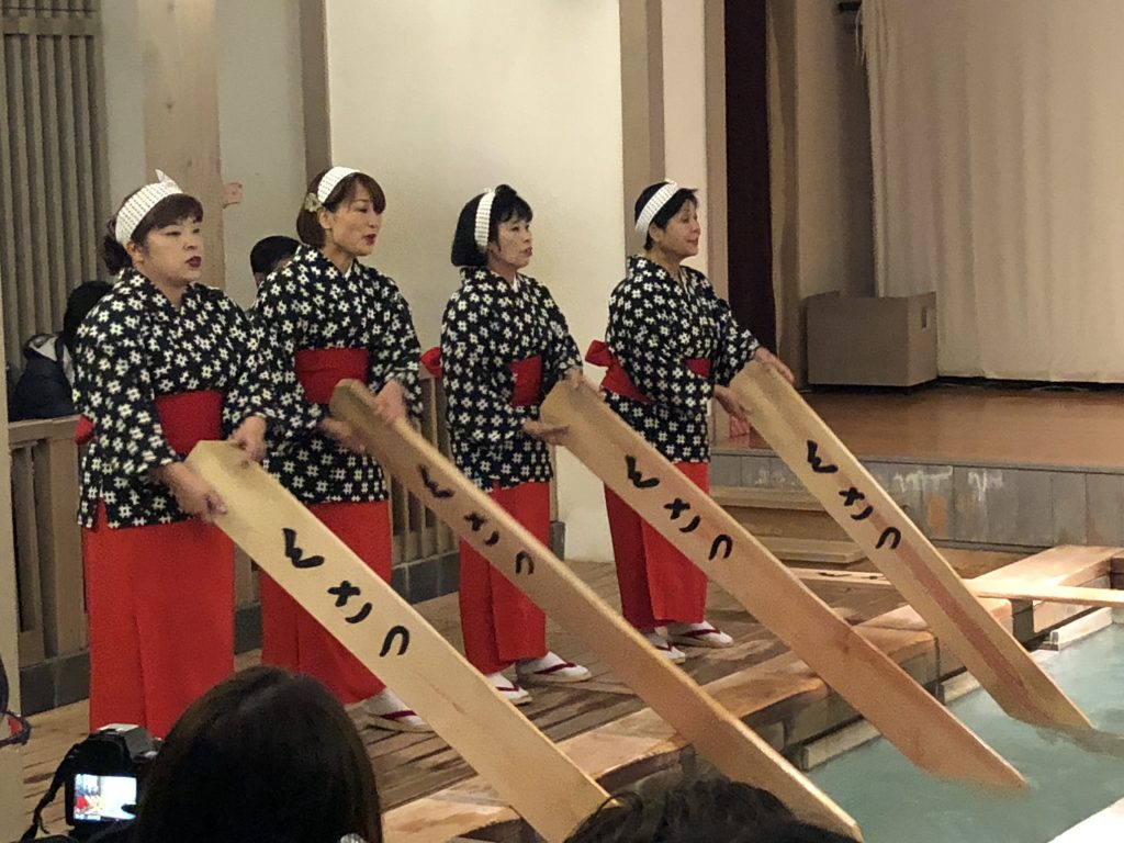 Four women in traditional garb with long wooden paddles that are dipped into the hot water. 