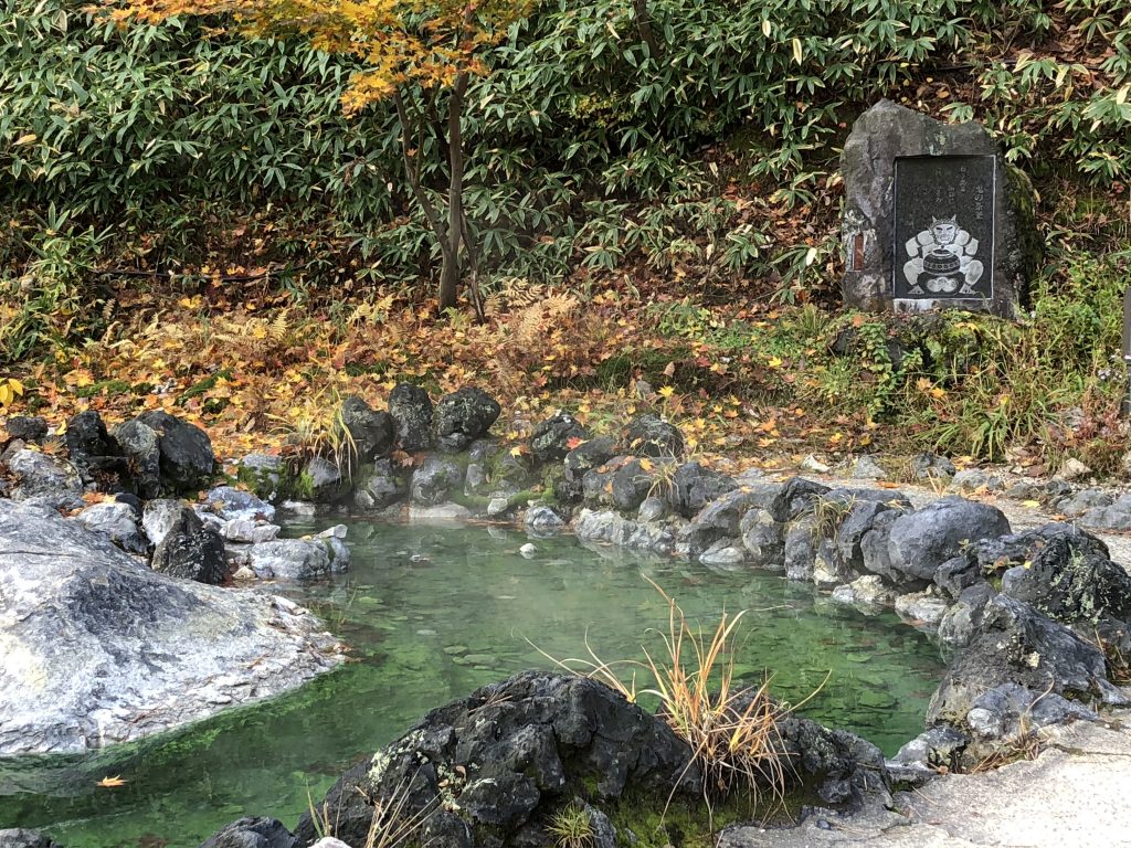 A small pool ringed with stone. You can see steam rising from the pool. In the background, there is a carving of an oni/devil with a poem carved above it.