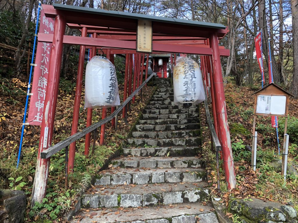 Steps leading up to a small Shinto shrine on a wooded hill. Red wooden torii gates line the stairs, and lanterns hang from them.