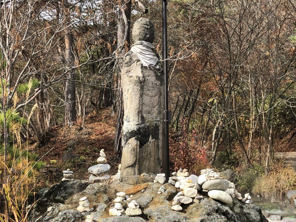 A very, very old statue. Most of its features have been worn away, but the tattered remains of a bib can still be seen around its neck, identifying it as Jizo. Small stacks of rocks have been left around it by adherents.