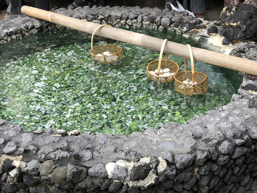 A long bamboo rod is suspended over a rock pool full of hot spring water. Three baskets full of eggs are hanging from the rod.