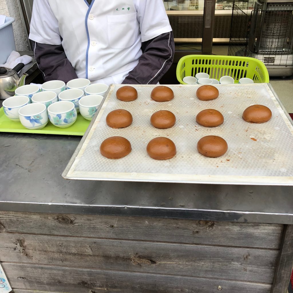 A close-up of the counter at a manjuu stand. There is a tray full of small brown manjuu and a tray full of empty teacups.