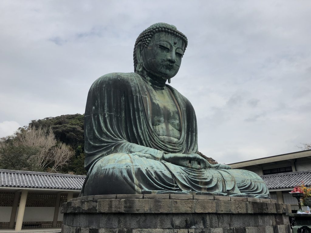 A photo of the great Buddha of Kamakura. It is a seated bronze statue that has a lovely patina. He is sitting in the lotus position.